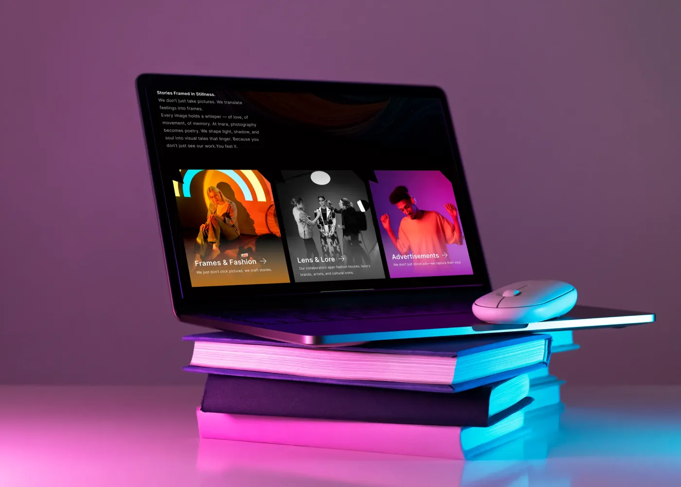 Laptop displaying a modern, dark-themed photography website layout, resting on a stack of books under vibrant pink and blue neon lighting.