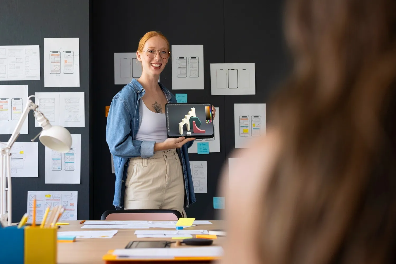 A designer presenting a premium uiux design on a tablet during a team meeting, with mobile wireframe sketches pinned to the wall behind her.