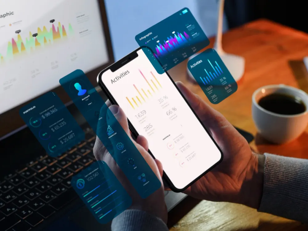 Hand holding a smartphone showing an analytics dashboard with activity charts and floating UI data cards, with a laptop and coffee cup in the background.